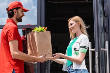 Smiling courier giving shopping bag with vegetables to young woman near building on urban street  clipart