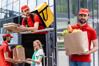 Gülümseyen kurye kolajı pizza kutularını tutuyor ve alışveriş torbalarını şehir sokaklarında kadınlara veriyor. 