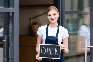 Smiling waitress holding signboard with open lettering near door of cafe on urban street  clipart
