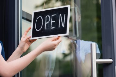 Cropped view of waitress hanging signboard with open lettering on door of cafe  clipart