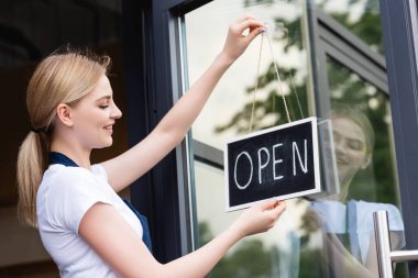 Side view of smiling waitress in apron hanging signboard with open lettering on door of cafe  clipart