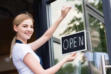 Cafe kapısında açık harflerle tabelayı asarken kameraya gülümseyen garsonun yan görüntüsü 