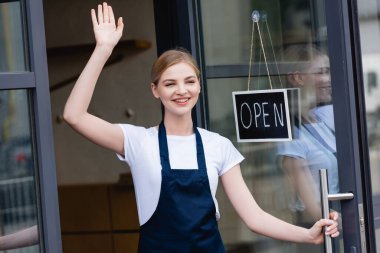 Positive waitress waving hand near signboard with open lettering on door of cafe  clipart