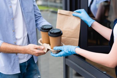 Cropped view of man receiving package and paper cups from waitress in latex gloves near entry of cafe  clipart