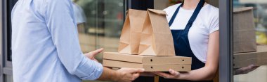 Panoramic shot of man receiving pizza boxes and paper bags for takeaway from waitress near window of cafe clipart