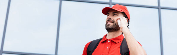Panoramic crop of smiling courier in uniform talking on smartphone on urban street 