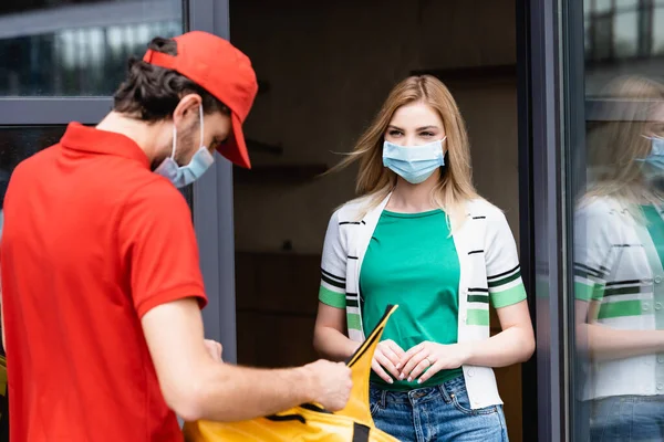 Tıbbi maskeli bir kadının şehir sokağında termal çantalı kuryenin yanında durmasının seçici odağı. 