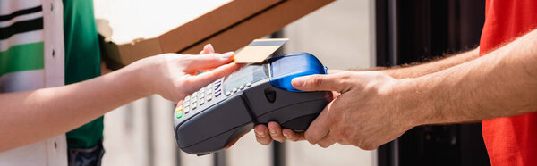 Panoramic crop of woman with pizza boxes holding credit card near waiter with payment terminal on urban street 