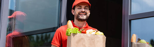Panoramic shot of positive delivery man holding shopping bag with grocery on urban street 