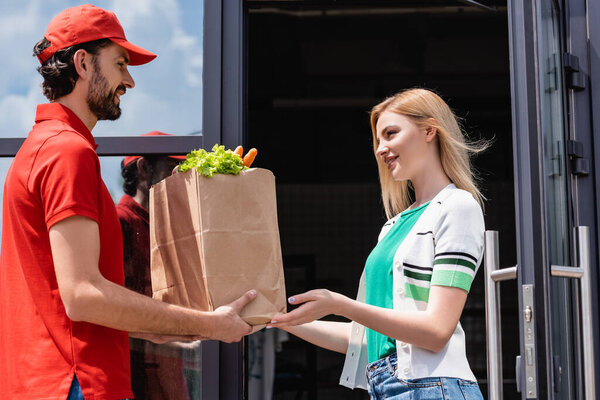 Smiling courier giving shopping bag with vegetables to young woman near building on urban street 