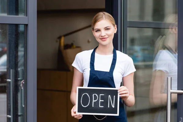 Smiling waitress holding signboard with open lettering near door of cafe on urban street