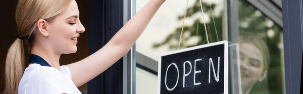 Panoramic shot of smiling waitress hanging signboard with open lettering on door of cafe 