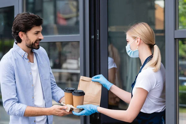 Side view of man receiving package and paper cups from waitress in latex gloves and medical mask near cafe