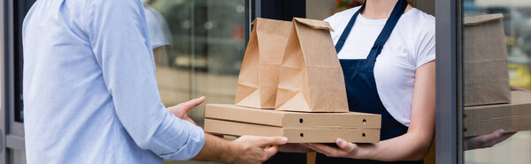 Panoramic shot of man receiving pizza boxes and paper bags for takeaway from waitress near window of cafe