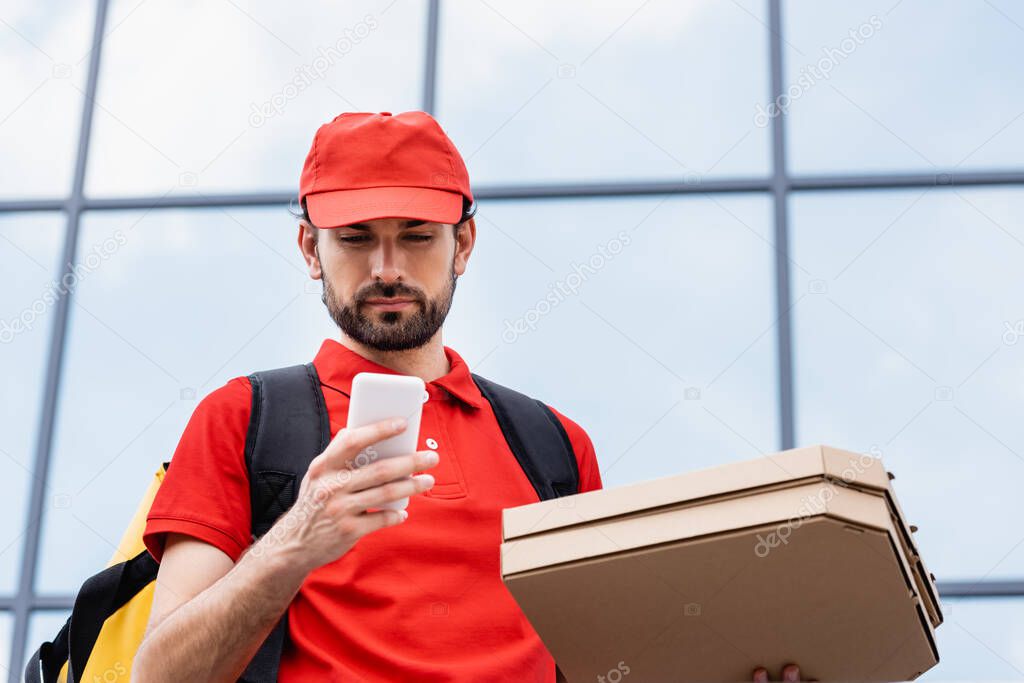 Low angle view of courier holding pizza boxes and using smartphone on urban street 
