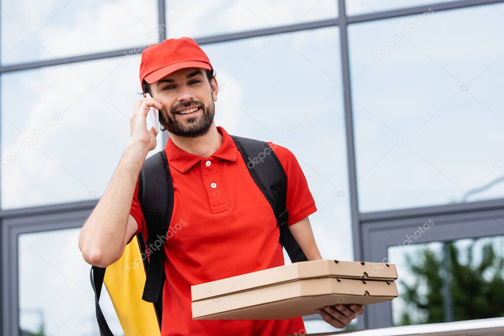 Smiling delivery man looking at camera while holding pizza boxes and talking on smartphone on urban street 
