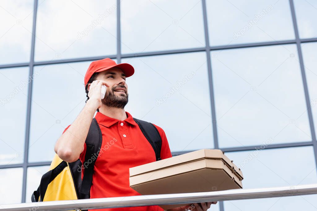 Low angle view of smiling courier talking on smartphone while holding pizza boxes on urban street 