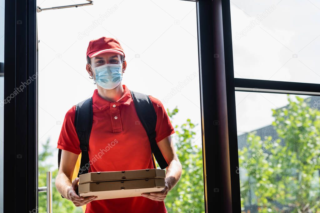Courier in medical mask holding pizza boxes near open door of building 
