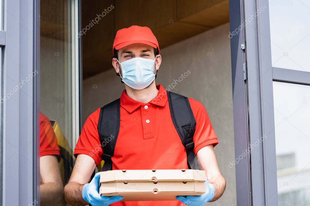 Delivery man in medical mask and latex gloves holding pizza boxes near building on urban street 