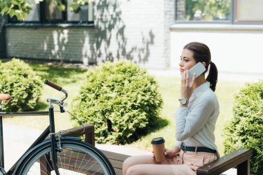 Güzel iş kadını akıllı telefondan konuşuyor ve bisikletin yanında kağıt bardak tutuyor. 