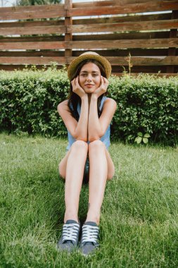 Hasır şapkalı neşeli genç kadın yüzüne dokunuyor ve çimenlerde otururken kameraya bakıyor.