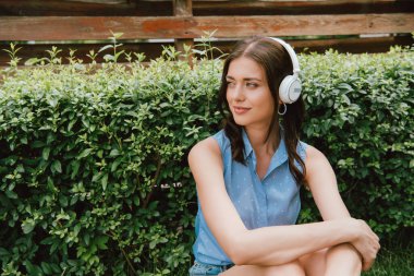 Neşeli genç bir kadın çalılığın yanındaki kablosuz kulaklıkla müzik dinliyor. 
