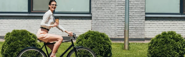 身穿眼镜 骑自行车 拿着纸杯的快乐女商人的全景照片 — 图库照片