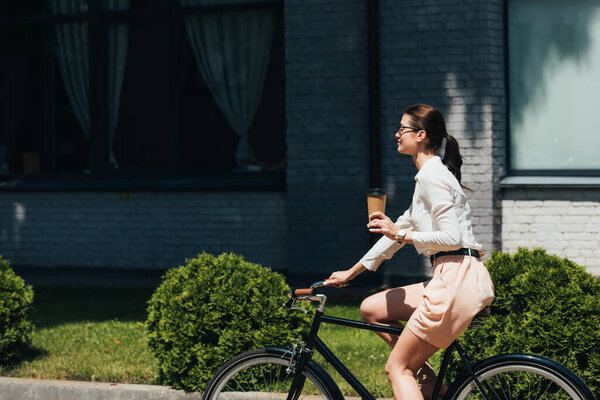 side view of happy businesswoman in glasses riding bicycle and holding paper cup