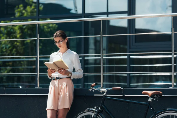 戴眼镜的漂亮女商人在自行车和现代建筑附近看书 — 图库照片