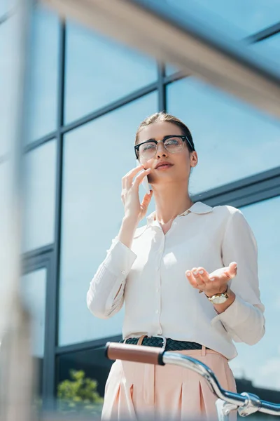 Selective Focus Attractive Businesswoman Glasses Talking Smartphone Gesturing Building Bicycle — Stock Photo, Image