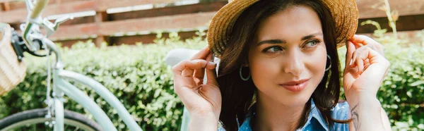 Bisikletin yanında hasır şapkaya dokunan çekici kadının yatay görüntüsü 