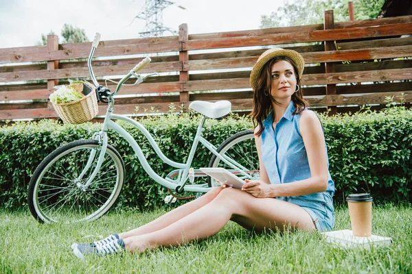 デジタルタブレットを持ち自転車や紙コップや本の近くに草の上に座っている藁帽子の女性 — ストック写真