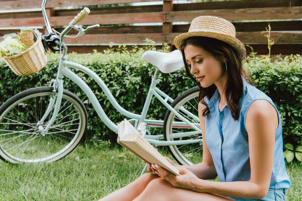 стильная девушка в соломенной шляпе читает книгу и сидит на траве возле велосипеда
