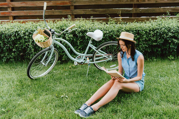 trendy girl in straw hat reading book and sitting on grass near bicycle with wicker basket 