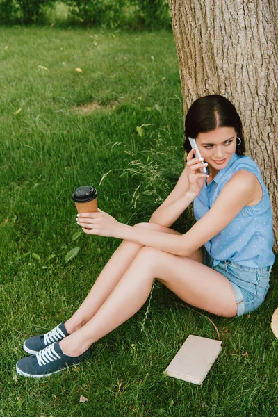 スマートフォンで話す魅力的な女性と草の上に座って紙コップを持ち — ストック写真