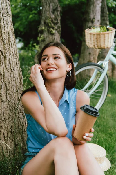 Uśmiechnięta Kobieta Rozmawiająca Smartfonie Trzymająca Papierowy Kubek Siedząc Pobliżu Pnia — Zdjęcie stockowe