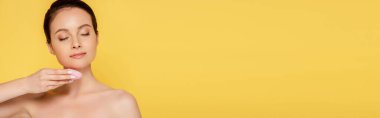 beautiful naked woman using facial cleansing brush with closed eyes isolated on yellow, panoramic shot clipart