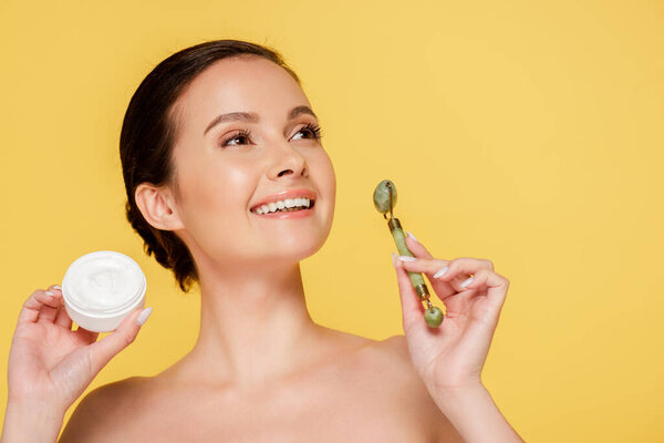улыбающаяся обнаженная красивая женщина с косметическим кремом и нефритовым роликом, изолированным на желтом