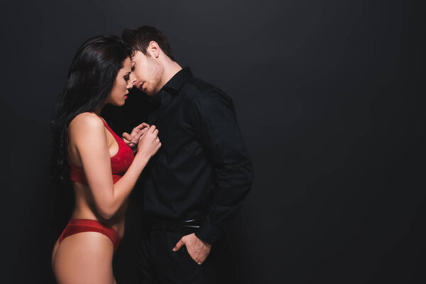 seductive woman in red underwear touching shirt of passionate boyfriend standing with hand in pocket isolated on black 