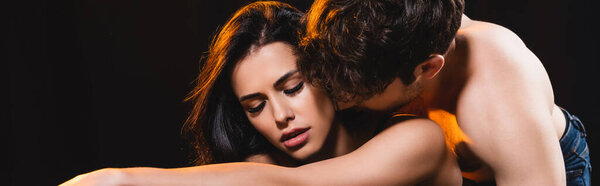 panoramic crop of muscular man kissing woman with closed eyes isolated on black 
