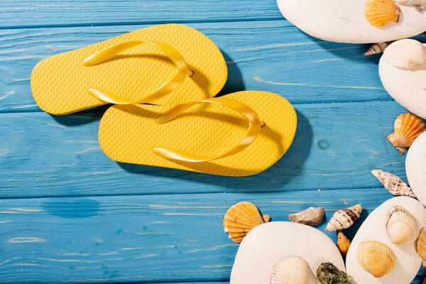 top view of yellow flip flops near seashells and stones on wooden blue background