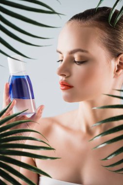 Makyajlı çekici kadın beyaz palmiye yapraklarının yanındaki toner şişesine bakıyor.