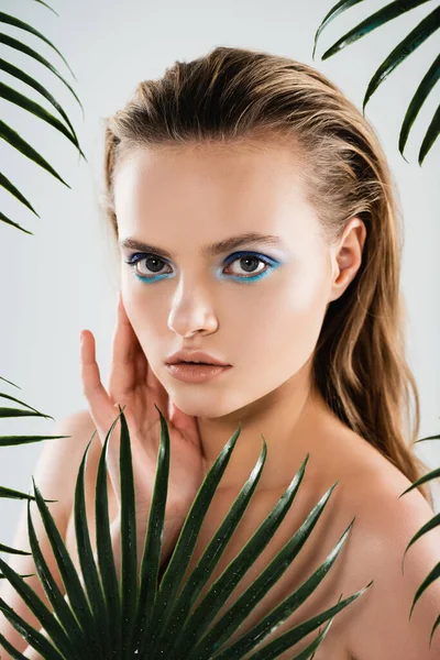 beautiful woman with makeup looking at camera near palm leaves on white