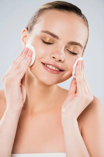 cheerful woman holding cotton pads near clean face isolated on grey