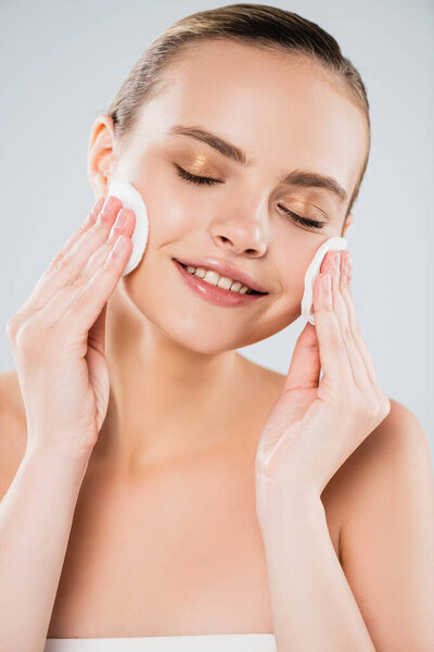 cheerful woman holding cotton pads near clean face isolated on grey 