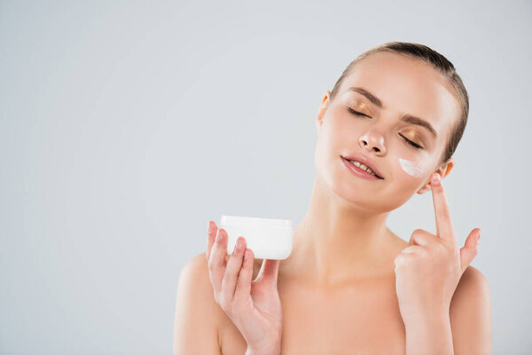 happy woman holding container while applying face cream isolated on grey 
