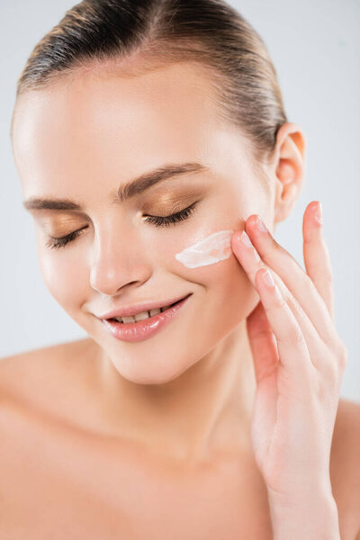 cheerful woman with closed eyes applying face cream isolated on grey 