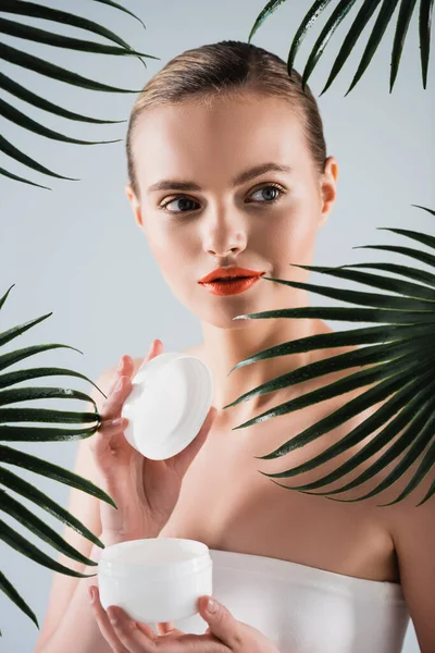 白い掌の葉の近くに顔クリームの入った化粧容器を持つ魅力的な女の子 — ストック写真