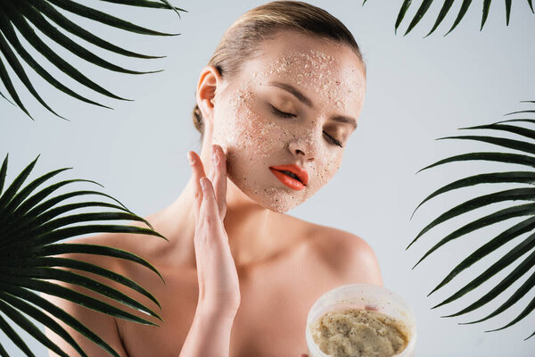 young naked woman applying scrub and holding container near palm leaves on white