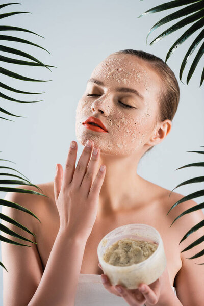 young woman applying scrub and holding container near palm leaves on white